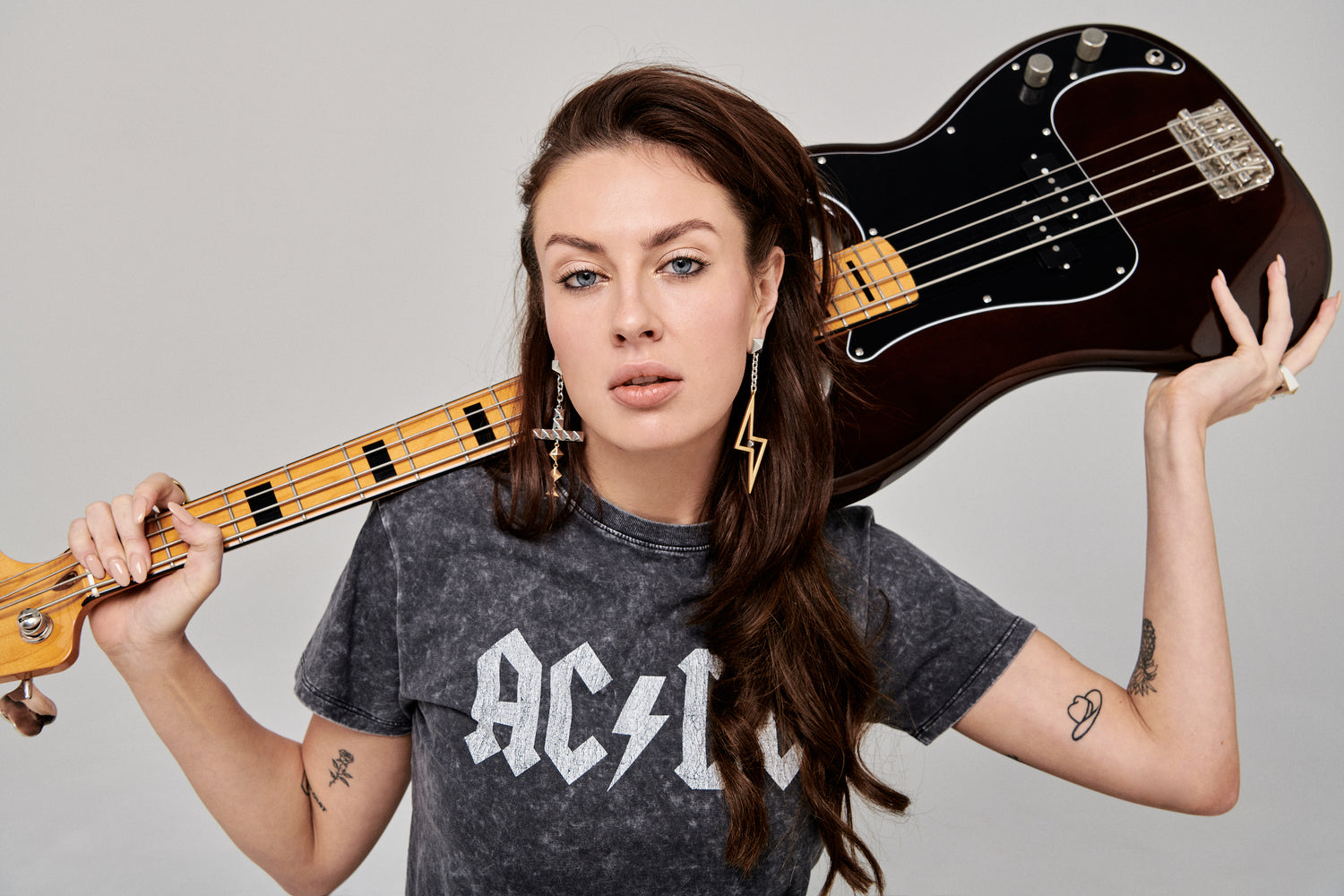 Model stands with bass guitar behind her head. Glamrock look with statement earring featuring mixed metals of sterling silver and 18k gold plating are framed by the bass. Asymmetrical styling with ACDC band tee and statement earrings Fame and New Romance.